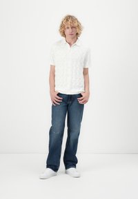 Wit kortom shirt met geperforeerd patroon, gecombineerd met blauwe recht gesneden jeans en witte sneakers; model staat met handen op de heupen.