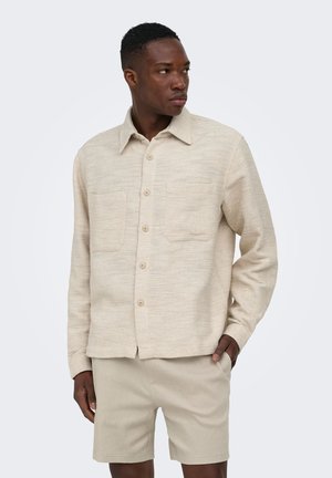 Homme portant une chemise beige texturée à boutons avec deux poches poitrine et un short beige assorti, regardant vers sa droite.