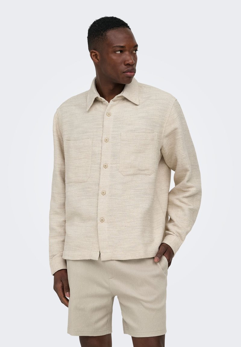 Homme portant une chemise beige texturée à boutons avec deux poches poitrine et un short beige assorti, regardant vers sa droite.