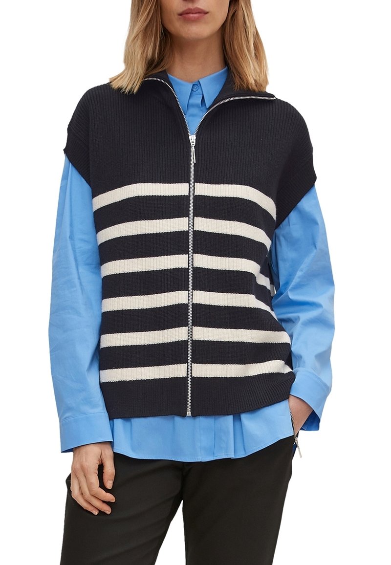 Femme portant un pull sans manches zippé rayé noir et blanc par-dessus une chemise bleu clair boutonnée avec un pantalon noir.