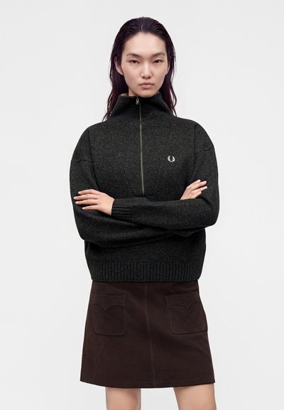 Jeune femme aux longs cheveux foncés portant un pull zippé gris foncé et une jupe marron foncé avec des poches avant, debout les bras croisés.