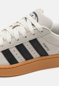 Calzado con parte superior de ante gris claro, diseño de tres rayas negro, cordones crema y suela de goma. Logo impreso en el lateral.