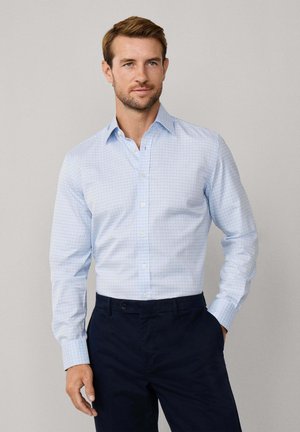 Homme portant une chemise de costume à carreaux bleu clair rentrée dans un pantalon noir marine, se tenant avec une main dans la poche devant un fond uni.