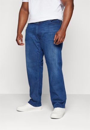 Homme portant un jean droit bleu, des baskets blanches et un t-shirt blanc, debout devant un fond clair uni.