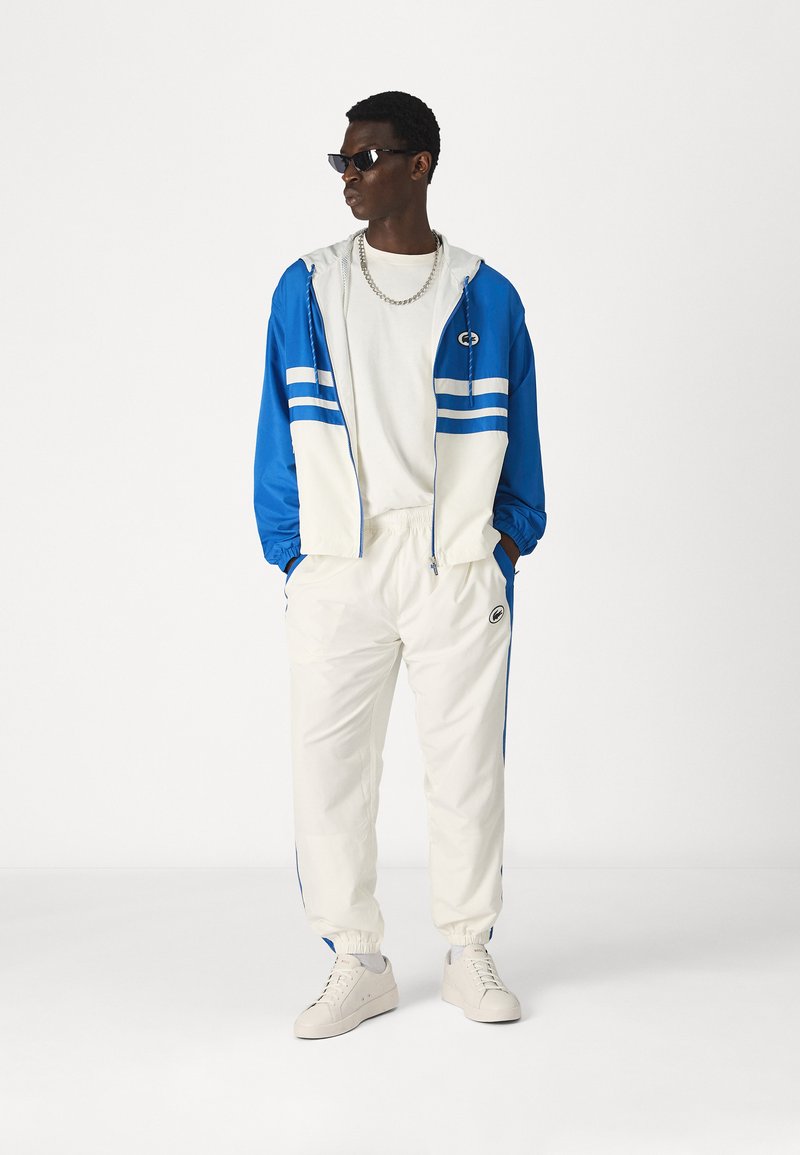 Survêtement bleu et blanc comprenant une veste zippée avec des rayures, un pantalon à taille élastique et des chaussures de sport, sur fond uni.