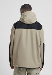 Giacca impermeabile beige con una parte superiore nera, dotata di cappuccio e maniche lunghe. Design dalla texture liscia, con hardware minimal e blocchi di colore a contrasto.