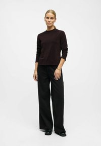 Vrouw in een donkere lange mouwen top, wijde zwarte broek en zwarte schoenen, staand tegen een eenvoudige witte achtergrond.