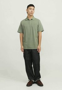 Πράσινο πόλο μπλούζα με γιακά, κοντά μανίκια και λογότυπο. Συνδυασμένη με σκούρο τζιν παντελόνι και καφέ παπούτσια, με απλό φόντο.