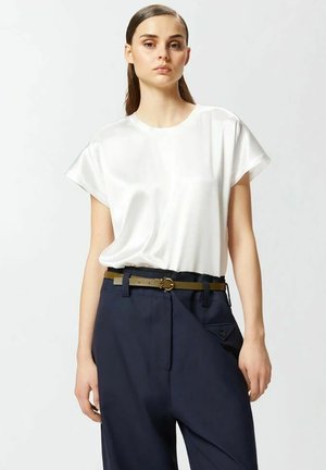 Giovane donna con capelli tirati indietro che indossa una camicetta bianca di raso a maniche corte infilata in pantaloni navy su misura con cintura sottile.