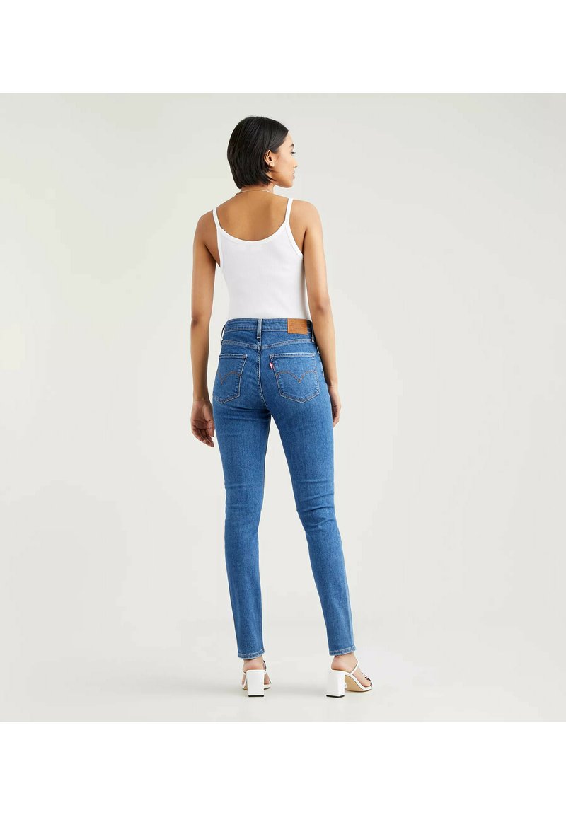 Schmale blaue Jeans aus Denim mit einer mittelhohen Taille und Gesäßtaschen. Ein weißes Tanktop und helle Absätze runden das Outfit ab.