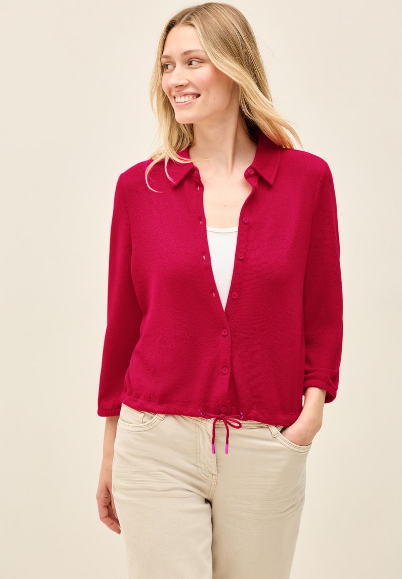 Magenta Strickjacke mit Knopfverschluss, Dreiviertelärmeln und verstellbarem Kordelzug am Saum. Getragen über einem weißen Oberteil und beigen Hosen.