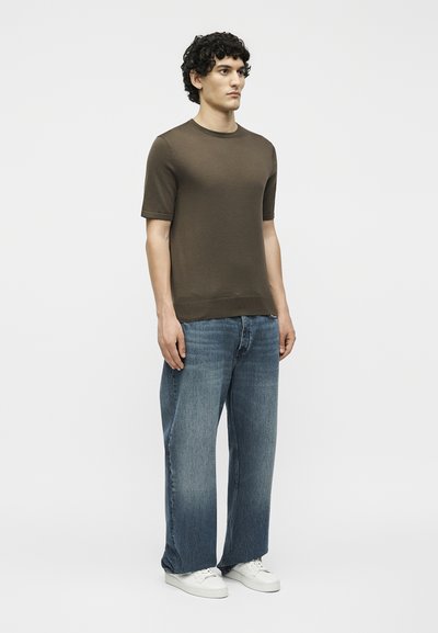 Άνδρας που φοράει καφέ κοντομάνικη πλεκτή μπλούζα, φαρδιά μπλε τζιν και λευκά αθλητικά παπούτσια μπροστά σε απλό ανοιχτό φόντο.