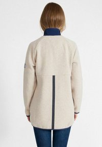 Beige Fleecejacke mit navyfarbenem Kragen und schwarzen Akzenten entlang des Rückens. Mit Raglanärmeln und geradem Saum.
