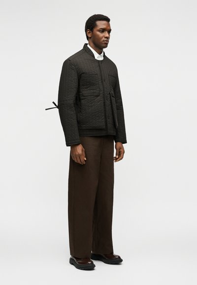 Homme portant une veste matelassée noire, une chemise blanche, un pantalon large marron et des chaussures marron foncé, debout devant un fond clair uni.