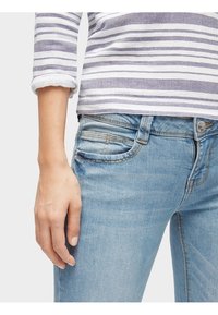 Vaqueros de mezclilla azul con un corte ajustado, diseño de cinco bolsillos y lavado claro. Parte de una blusa de rayas de manga larga con líneas grises y blancas es visible.