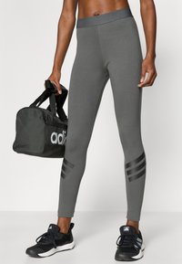 Sivé legíny s priliehavým strihom, vysokým pásom a troma čiernymi pruhmi na dolných častiach nôh. Doplnkové čierne športové taška a topánky.