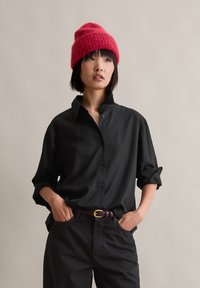 Svart skjorte med knapper og avslappet passform, med oppbrettede ermer og krage, kombinert med høy midje mørke bukser og en rød strikket lue.