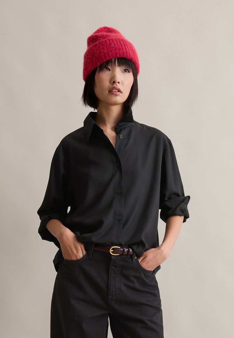 Svart skjorte med knapper og avslappet passform, med oppbrettede ermer og krage, kombinert med høy midje mørke bukser og en rød strikket lue.