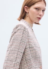 Veste tissée en rose pastel et blanc avec un motif texturé. Elle présente un col montant et une fermeture ronde à boutons.