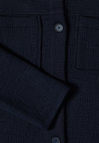 Manche et devant de veste texturée bleu marine foncé avec deux boutons et une poche avant visible sur un tissu à motif de quadrillage.