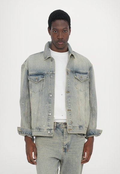 Denim jacket, light blue with slight distressing, two chest pockets, silver buttons, and decorative stitching. Paired with a white T-shirt.
