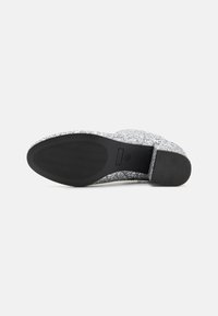 Zapato plateado brillante tipo slip-on con una superficie texturizada, suela plana negra y puntera redondeada. Presenta sutiles acentos negros en el talón.