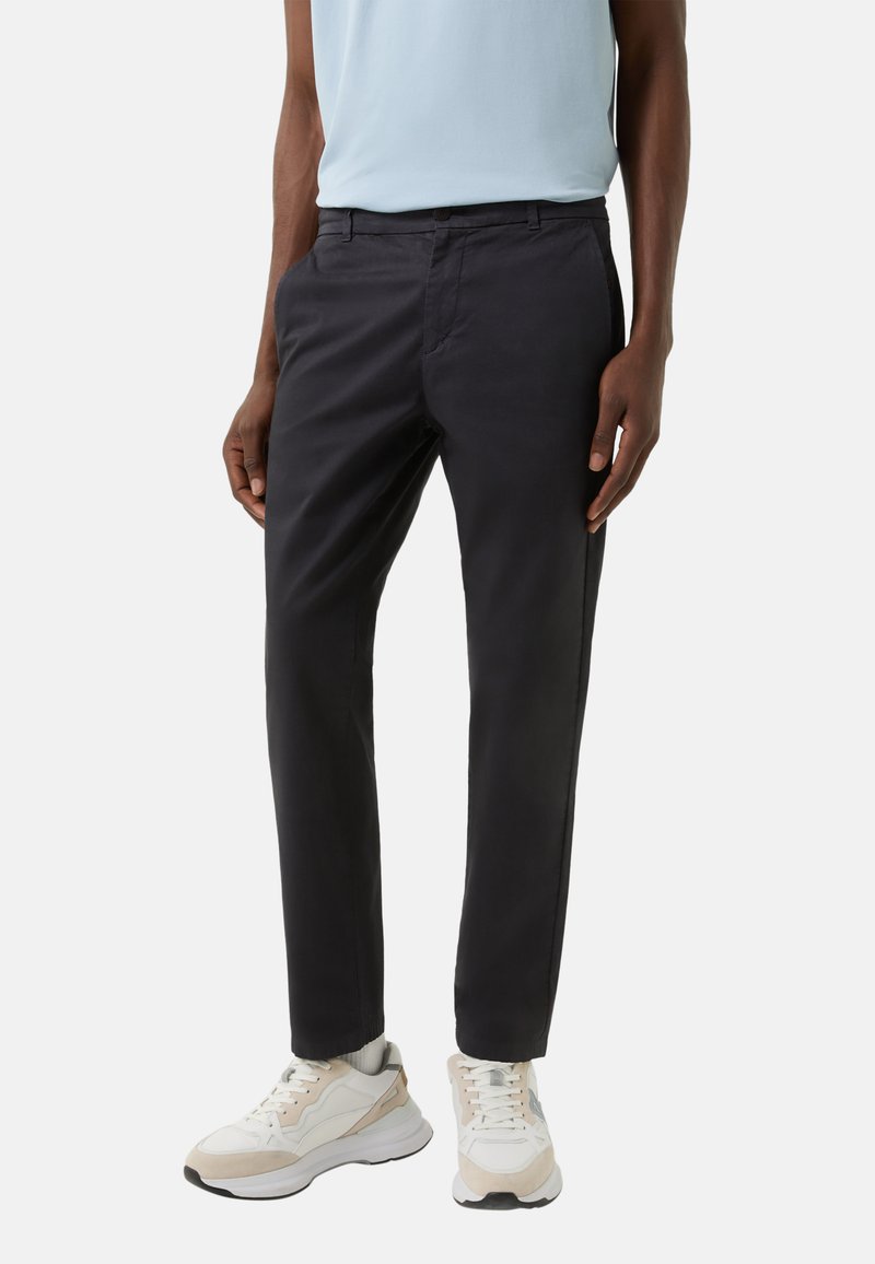 Schwarze, zulaufende Hose mit glatter Textur, ausgestattet mit einem Knopfverschluss und Seitentaschen, kombiniert mit weißen und beigen Sportschuhen.