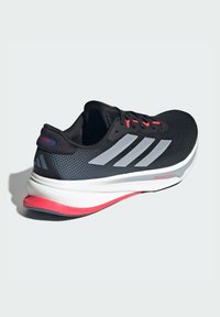 Zapatilla de running negra con parte superior de malla, tres franjas plateadas, una entresuela blanca y acentos rojos en la suela. Cuenta con cierre de cordones y un cuello acolchado.