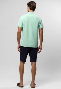 Mężczyzna stojący tyłem, ubrany w jasnozieloną koszulkę polo z krótkim rękawem, czarne szorty i brązowe klapki na jednolitym tle.