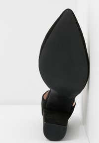 Escarpin noir en daim à talon haut avec un orteil pointu et une semelle texturée, présentant une finition douce et un design élégant et minimaliste.