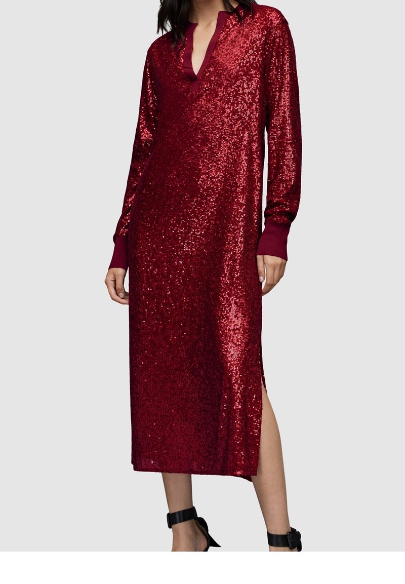 Femme portant une robe longue à manches longues, rouge foncé pailletée, avec un décolleté en V et une fente sur le côté, associée à des talons noirs à bride à la cheville.