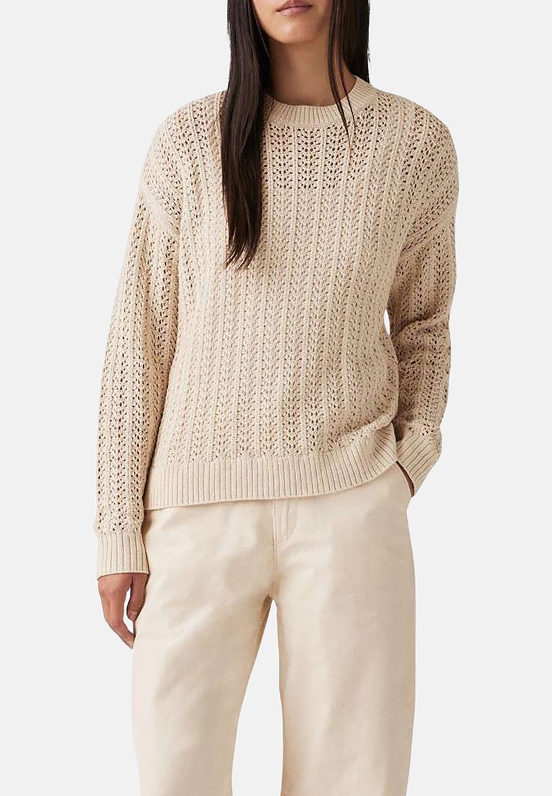 Beige gestrickter Pullover mit ribbeliger Textur und offenem Stichmuster, runder Ausschnitt und langen Ärmeln. Kombiniert mit hellen Hosen.