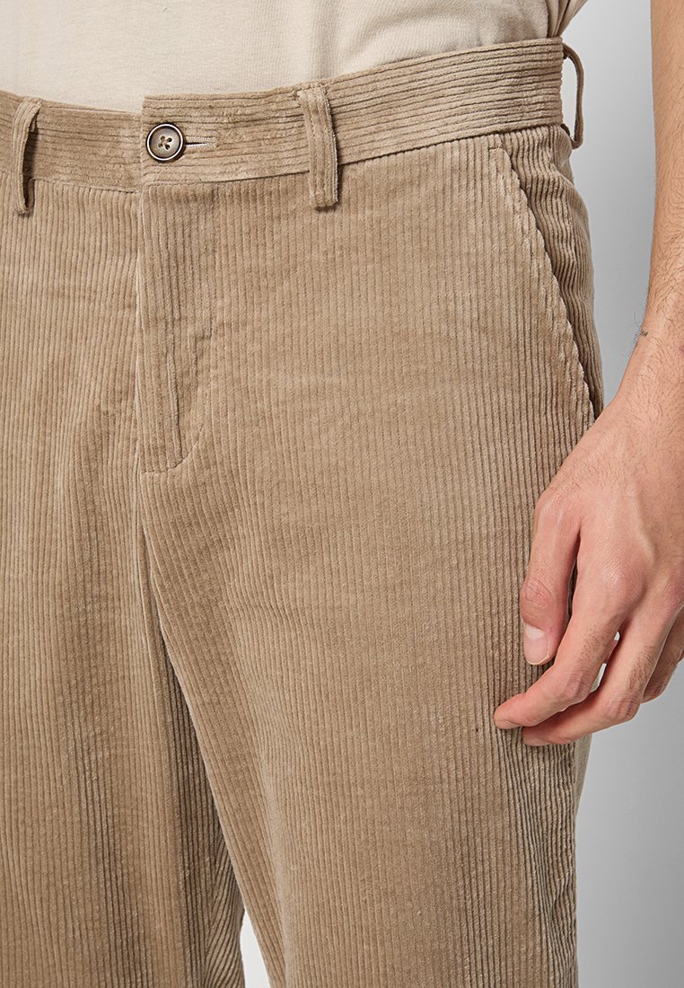 Beige Cord-Hose mit vertikalen Rippen, vorn Knopf, Gürtelschlaufen, Seitentasche und einer rechten Hand, die nahe am Oberschenkel ruht.