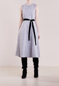 Robe grise sans manches à silhouette évasée, avec une ceinture en velours noir à la taille, portée avec des bottes noires montantes jusqu'aux genoux.
