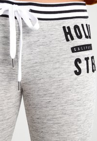 Šedé tepláky s texturovaným povrchem, s černobílým pruhovaným pasem, černým potiskem a bílou šňůrkou na stažení.