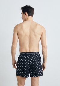 Čierne boxerky s opakujúcim sa bielym vzorom a čiernym elastickým pásom s textom „KARL LAGERFELD“. Mäkko materiál, voľný strih.
