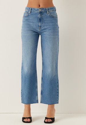 Lichtblauwe high-waisted rechte spijkerbroek met een onafgewerkte zoom, blootsvoets gedragen met zwarte open teen pumps met strikjes aan de voorkant.