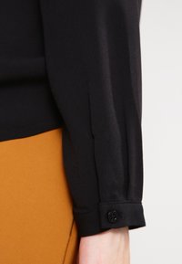 Schwarzes Langarmshirt mit glatter Textur, ausgestattet mit einem Knopf am Ärmelbund und einem entspannten Schnitt, kombiniert mit einem braunen Stoff darunter.
