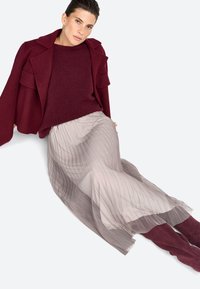 Maglione in lana bordeaux abbinato a una gonna plissettata di un leggero color taupe. L'outfit presenta un giacca strutturata e aperta sul davanti e stivali al ginocchio.