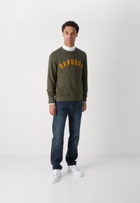 Olivově zelená mikina s oranžovým nápisem "BARBOUR", spárovaná s tmavě modrými džíny a bílými teniskami. Hladká tkanina a uvolněný střih.