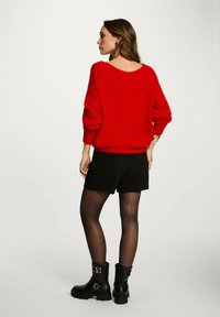 Maglione rosso a maglia con scollo ampio, pantaloni corti neri, collant neri trasparenti e stivali neri con lacci e dettagli in oro.