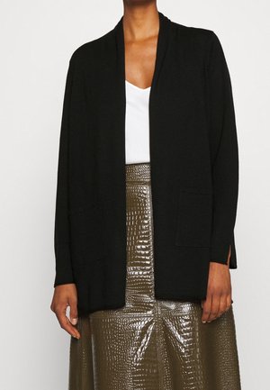 Personne portant un cardigan noir ouvert sur un top blanc et une jupe brillante vert olive à texture crocodile.