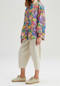 Kobieta w beżowych, krótkich szerokich spodniach, beżowych sneakersach oraz luźnej, żółtej koszuli w kwiaty z fioletowymi, różowymi i niebieskimi kwiatami.