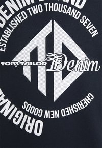 Texte blanc et logo sur tissu foncé : « Tom Tailor Denim », « Fondé en deux mille sept », « Original » et « Nouveaux produits précieux ».