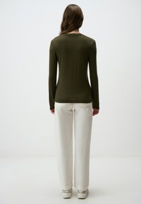 Maglietta a maniche lunghe verde oliva con vestibilità slim, tessuto morbido e scollatura a girocollo, abbinata a pantaloni bianchi a gamba larga e sneakers di colore chiaro.