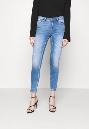 Jeans ajustados azules con detalles desgastados, cintura de media altura y un tono desgastado. Presenta cremalleras en los tobillos y diseño de cinco bolsillos.