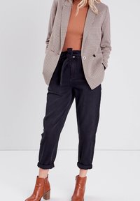 Kvinna iklädd en brun polotröja, rutigt blazer, svarta högmidjade upprullade byxor med bälte och bruna ankelstövlar.