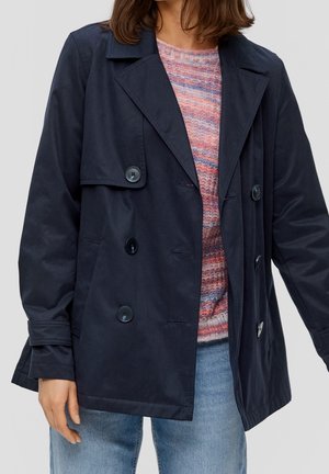 Mujer con un abrigo tipo gabardina azul marino de doble botonadura sobre un suéter de rayas rosas y moradas y jeans azul claro, de pie contra un fondo liso.