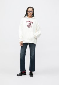 Kremna majica s kapuco z bordo napisom "TOMMY 85 AUTHENTIC ORIGINAL", kombinirana s temno modrimi razširjenimi kavbojkami in črnimi lakastimi čevlji.