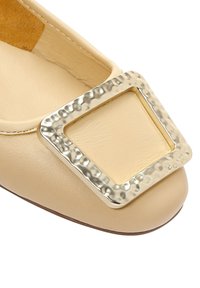 Scarpa piatta in pelle beige con una grande fibbia in oro spazzolato sulla punta e dettagli cuciti lungo i bordi.